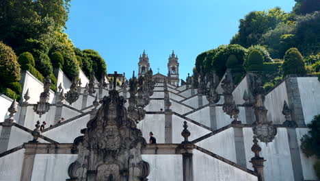 Bottom-up-shot-of-tourist-walking-up-Baroque-stairway-to-famous-Bom-jesus-do-Monte-in-Braga-against-blue-sky---Slow-motion-wide-shot