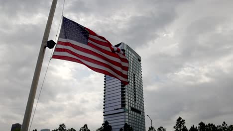 Im-Wind-Wehende-Amerikanische-Flagge-Mit-Einem-Gebäude-Im-Hintergrund-In-Houston,-Texas,-Mit-Stabilem-Video