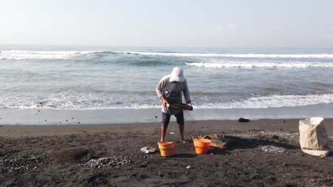 Trabajador-Recoge-Rocas-Negras,-Playa-En-Bali-Indonesia,-Técnica-De-Trabajo-Tradicional
