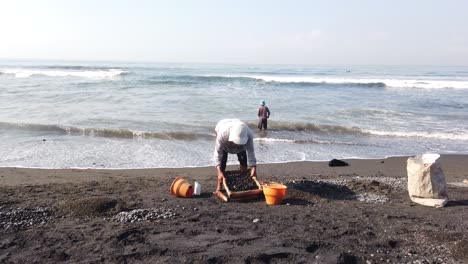 Arbeiter-Sammeln-Schwarze,-Abgerundete-Steine-Am-Strand,-Traditionelle-Arbeit,-Bali,-Indonesien