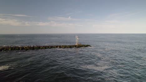 Aerial-drone-shot-pan-around-light-house-bouy-on-rock-jetty-as-man-fishes-with-heavy-lens-flare