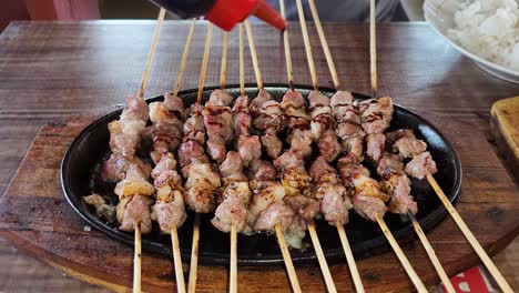 Freshly-cooked-goat-satay-on-hot-plate-being-topped-with-soy-sauce,-close-up