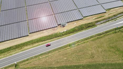 Aerial-isolated-electric-red-car-driving-along-asphalted-road-with-solar-panel-photovoltaic-power-plant-during-a-sunny-day,-rechargeable-lithium-battery-zero-emission-alternative-sustainable-energy