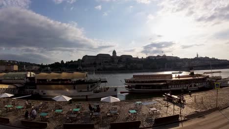 Barcos-Atracados-En-El-Muelle-De-Budapest-Durante-La-Puesta-De-Sol,-El-Río-Danubio,-El-Castillo-De-Buda,-El-Bastión-De-Los-Pescadores-Y-El-Monasterio-Carmelita-Al-Fondo,-Cámara-Lenta---Budapest,-Junio-De-2023