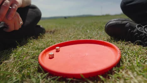 Two-people-sitting-on-grass-and-throwing-dice-onto-orange-disc,-slow-mo