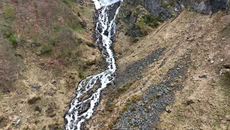 Small-waterfall-at-Grasteindalselva-grey-stone-valley-river-in-a-Geirangerfjord-mountain-close-to-ornevegen-eagles-road---Spring-aerial-with-tilt-up-during-melting-snow-and-yhellow-grass