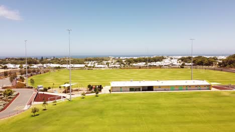 Sports-ground-with-building,-floodlights-with-blue-sky-and-ocean-background,-aerial-Perth-Australia
