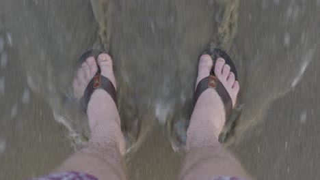 Men-feet-in-sandals-stand-on-the-beach---waves-come-easily-to-the-beach-and-cover-the-feet