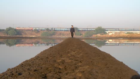 Mann-Läuft-An-Einem-Sonnigen-Morgen-Auf-Dem-Schlammigen-Gehweg-Zwischen-Teichen-In-Mumbai,-Indien,-Im-Hintergrund-Fährt-Ein-Nahverkehrszug-–-Totale