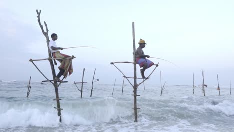 Pescadores-Sobre-Pilotes-Usando-Una-Caña-De-Pescar-Para-Pescar-Con-Olas-Chapoteando-En-La-Costa-De-Weligama,-Sri-Lanka