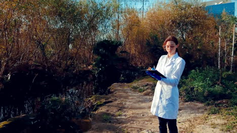 A-young-woman-scientist-at-a-creek,-wearing-protective-eyewear-and-a-lab-coat,-taking-notes-on-a-clipboard