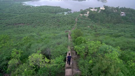 Männlicher-Tourist-Mit-Blick-Auf-Einen-Schmalen-Betonweg-Im-üppigen-Grünen-Tal,-Der-Zum-Wunderschönen-See-In-Udaipur,-Rajasthan,-Indien-Führt-–-Weitwinkelaufnahme