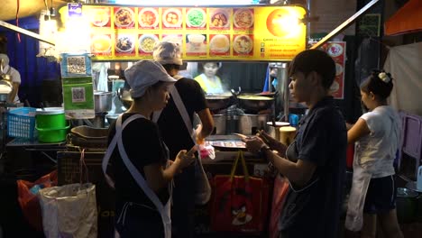 Ein-Mann-Zahlt-Einer-Verkäuferin-Geld-Für-Das-Essen,-Das-Er-Am-Imbissstand-In-Chinatown,-Bangkok,-Thailand-Gekauft-Hat-–-Tilt-up-Zeitlupenaufnahme