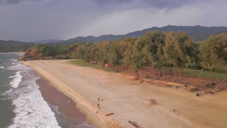 Drone-Fly-Towards-Crashing-Waves-With-Tourist-On-Fine-Sands-In-Dense-Trees-Foliage-At-Rajbagh-Beach,-Canacona-In-South-Goa,-India