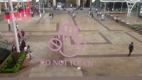 Do-Not-Touch-Hand-Icon-On-The-Glass-Wall-With-View-Of-People-Strolling-On-The-Ground-Of-A-Shopping-Mall-In-Mumbai,-India---close-up,-slow-motion