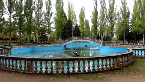 Empty-blue-pond-with-bridge-surrounded-by-trees-in-Virgen-de-las-Vinas-Park,-pan