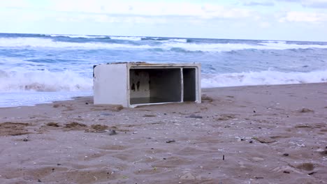 Old-fridge-washed-ashore-on-beach-with-sea-waves-crashing-in-background