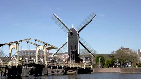 A-static-shot-of-the-historic-Molen-de-Put-windmill-beside-the-Rembrandt-bridge-as-people-go-about-their-day-on-a-beautiful-day-in-Leiden,-Netherlands