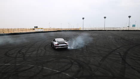 Aerial-tracking-shot-of-a-grey-Porsche-GT3-RS-doing-donuts-and-burnouts-in-an-arena