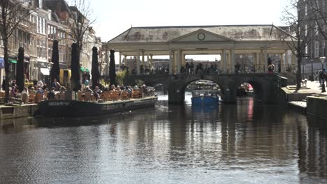 Barco-Pasando-Bajo-El-Puente-Koornbrug-Sobre-El-Río-Rin-Con-Gente-Disfrutando-Sentado-Afuera-Al-Lado-De-Un-Café-En-Lieden