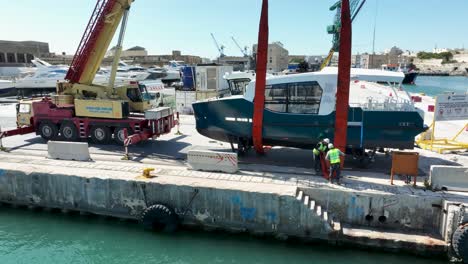 Toma-Aérea-De-Un-Dron-Orbitando-En-Sentido-Antihorario-Alrededor-De-Un-Barco-Levantado-En-El-Aire-Mientras-Los-Trabajadores-Colocan-Correas-De-Elevación-A-Su-Alrededor-En-El-Astillero-Mmh-De-Malta