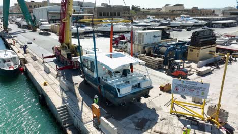 Toma-Aérea-De-Un-Dron-Orbitando-Lentamente-En-Sentido-Antihorario-Alrededor-De-Un-Barco-Levantado-En-El-Aire-Mientras-Lo-Mueve-Hacia-El-Mar-En-El-Astillero-Mmh-Malta