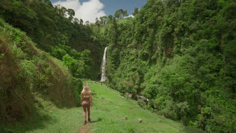 Woman-in-sportswear-hiking-downhill-towards-tropical-waterfall-in-lush-green-valley