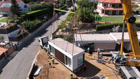 Aerial-view-construction-workers-checking-modular-housing-unit-suspended-from-heavy-crane-on-sunny-building-site