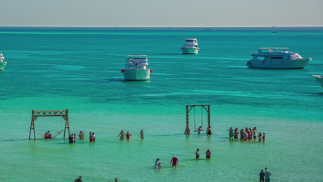 Timelapse-Tourists-waiting-for-beachside-swings-in-turquoise-water-as-boats-sail