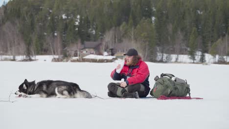 Rucksacktourist-Trinkt-Wasser,-Während-Er-Sich-Mit-Seinem-Alaskan-Malamute-Hund-In-Gefrorener-Landschaft-Ausruht