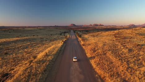 Vista-Aérea-De-Un-Automóvil-Blanco-Que-Conduce-A-Través-De-Un-Paisaje-De-Pradera-Iluminado-Por-El-Sol-En-Namibia