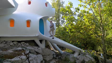 Man-Climbing-the-Steps-of-a-White-Rounded-House-in-the-Forest-and-open-the-door,-with-a-View-of-the-"Bubble-House"-and-Sun-Rays-Peeking-Through-the-Trees