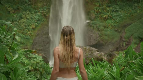 Stunning-European-blond-woman-in-white-bikini-walking-through-lush-vegetation-with-waterfall