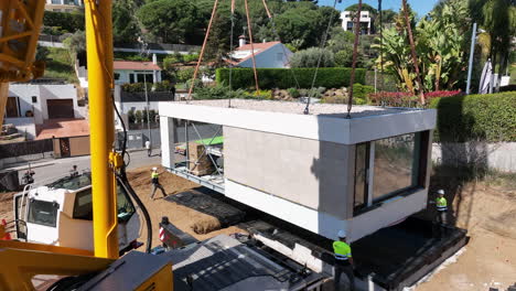 Aerial-view-of-construction-workers-guiding-smart-modular-home-onto-foundation-with-heavy-industrial-crane