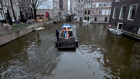 A-government-council-worker-standing-on-a-barge-cleaning-rubbish,-trash-and-litter-from-a-canal-in-Amsterdam,-Netherlands