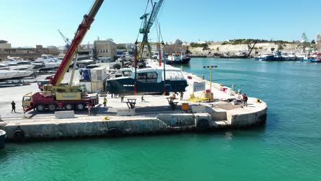 Drohnen-Luftaufnahme,-Die-Im-Uhrzeigersinn-Um-Ein-In-Die-Luft-Gehobenes-Boot-Kreist,-Während-Es-Auf-Der-MMH-Werft-In-Malta-Ins-Meer-Bewegt-Wird