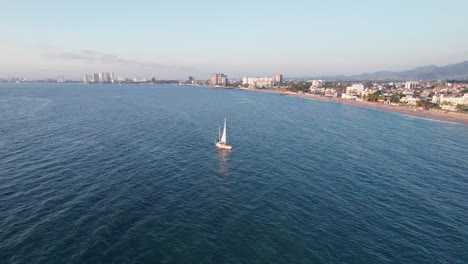 Drone-video-orbiting-around-a-sailboat-cruising-along-the-coast-of-Puerto-Vallarta,-showcasing-not-only-the-sailboat-but-also-the-coast,-mountains,-and-part-of-the-city