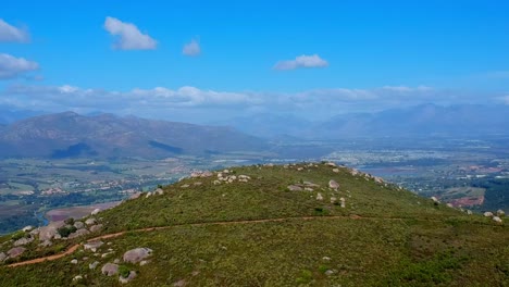 Vista-De-Arial-Drone-De-La-Cima-De-La-Colina-Con-Montañas-En-La-Distancia-Y-Casas-En-El-Valle-De-Abajo,-Paarl-Sudáfrica