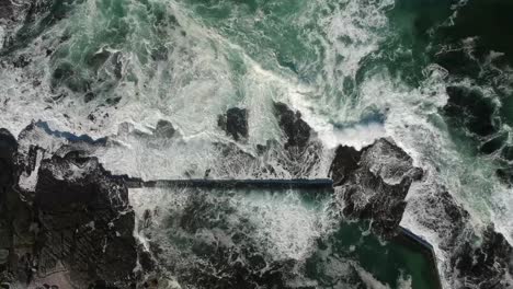 Rising-top-down-drone-view-of-tidal-pool,-with-white-water-waves-crashing-over-rocks