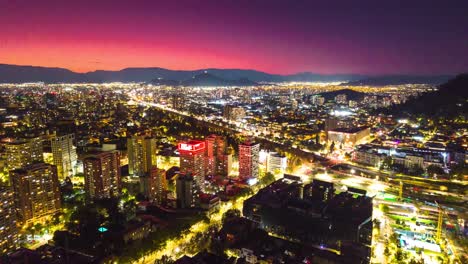 Aerial-Hyperlapse-View-of-Modern-Metropolitan-City-of-Santiago-Chile,-Baquedano-Neighborhood-Shining-at-Night,-Magenta-and-Gold-Andean-Cordillera-Background