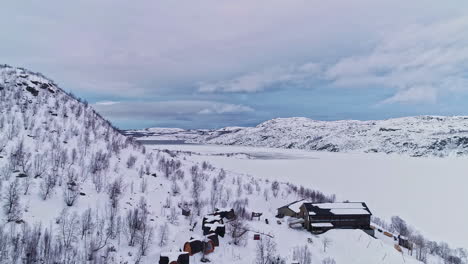 Aerial-backward-moving-cinematic-drone-shot-through-white-snowy-winter-valley-with-village-houses-surrounded-by-stunning-mountain-range-on-a-cloudy-day