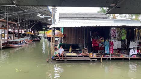 Vendedores-De-Ropa-Y-Souvenirs-Se-Sentaban-Junto-A-Sus-Puestos-Y-Esperaban-A-Los-Clientes-En-El-Mercado-Flotante-Damnoen-Saduak,-Provincia-De-Ratchaburi,-Al-Suroeste-De-Bangkok,-Tailandia