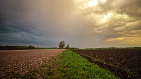 Shot-of-dark-cloud-movement-in-timelapse-over-ploughed-farmlands-on-both-sides-of-a-rural-path-during-evening-time