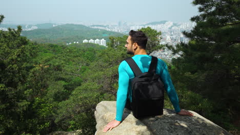 Hiker-Traveler-Man-Sitting-On-Steep-Cliff-Overlooking-Majestic-Urban-Seoul-City-Panorama-From-Gwanaksan-Mountain