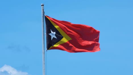 National-flag-of-Timor-Leste-flapping-in-breeze-against-tropical,-sunny-blue-sky-in-East-Timor,-Southeast-Asia
