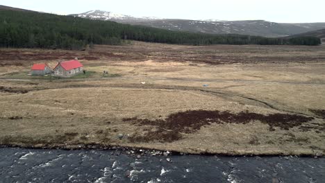 A-drone-hovers-over-a-river-facing-towards-a-pine-plantation-in-the-mountains-of-Scotland-and-a-remote-building-with-a-red-roof-next-to-4x4-track
