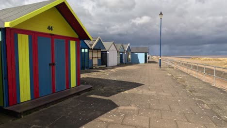 Colorful-beach-huts-stood-in-a-line-along-the-seafront-with-sandy-beach-and-moody-grey-sky’s