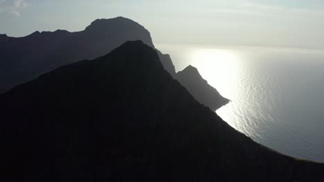 Drone-shot-of-Lofoten-steep-cliffs-and-mountains-sillhouetes-rising-from-deep-blue-sea