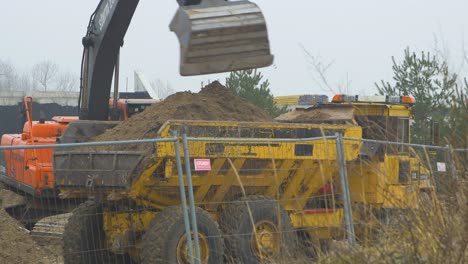 Intensive-work-on-the-construction-site-located-near-the-coast-of-the-sea,-loader-fills-the-truck,-white-sand-piles,-overcast-day-with-fog,-distant-medium-shot