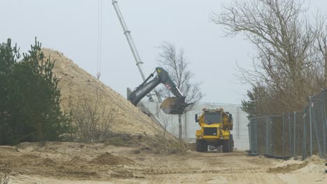Intensive-work-on-the-construction-site-located-near-the-coast-of-the-sea,-white-sand-piles,-loader-fills-the-truck,-overcast-day-with-fog,-distant-medium-shot